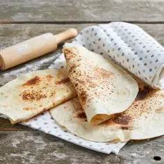 Placki do tortilli na drewnianym stole, obok wałek i ściereczka w kropki.